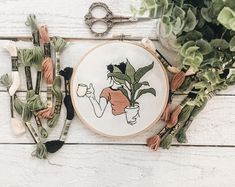 an embroidery project with scissors and flowers on a white wooden table next to some plants