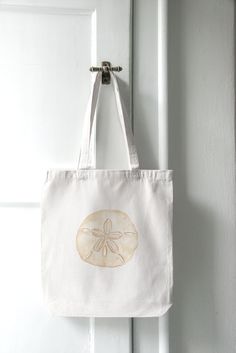 This coastal canvas tote bag is ready for a trip to the beach. Whether you're heading to the beach, strolling through a farmer's market, or simply running errands around town, this tote bag is your ideal companion. This Sand Dollar Cotton Canvas Tote Bag is the perfect blend of coastal charm with a trendy tan design.  🐚 Design: The Sand Dollar Tote Bag features a neutral watercolor painting that evokes the calmness of a morning by the beach. Its intricate sand dollar pattern, rendered in soft, earthy tones, adds a touch of coastal allure to your style, making it perfect for beach lovers and ocean enthusiasts. 🌞 Quality: Crafted from high-quality, durable cotton canvas, this tote bag is built to withstand the test of time. Rest assured that you can carry your essentials, from groceries an Casual White Beach Bag With Canvas Lining, Cream Cotton Beach Bag For Travel, Beige Cotton Canvas Bag For Vacation, Cream Cotton Bag For Vacation, White Beach Bag With Canvas Lining, White Beach Bag With Canvas Lining For Everyday, White Coastal Bags For Everyday Use, White Everyday Beach Bag With Canvas Lining, Cream Cotton Beach Bag For Vacation