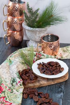 some pecans are sitting on a table next to two copper mugs with pine needles in them