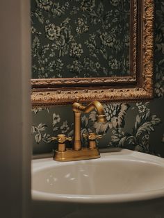 a bathroom sink with a gold faucet in front of a floral wallpaper