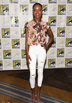 a woman standing in front of a wall wearing white jeans and a floral shirt with her hands on her hips