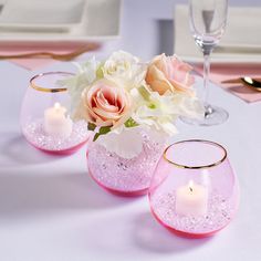 three pink vases with flowers and candles on a white table cloth, one is filled with water