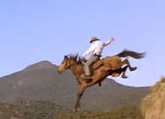 a man riding on the back of a brown horse over a grass covered hillside with mountains in the background