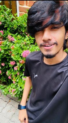 a man with black hair and a goatee standing in front of some pink flowers