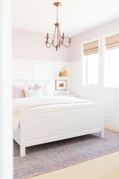 a white bed sitting under a chandelier in a bedroom next to a window