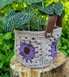 a crocheted basket with purple flowers and leaves in it sitting on top of a tree stump