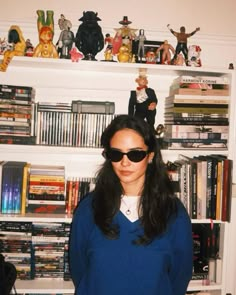 a woman wearing sunglasses standing in front of a book shelf filled with toy figurines