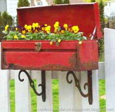 a red box with flowers in it sitting on a white picket fence next to green grass