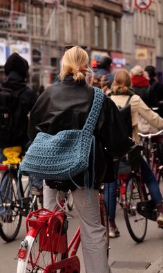 a woman walking down the street with a crocheted bag on her back and a red bicycle behind her