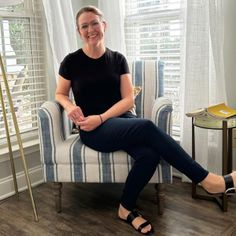 a woman sitting on a chair in front of a window smiling at the camera with her legs crossed