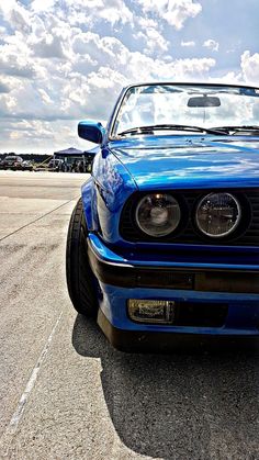 the front end of a blue sports car parked in a parking lot with clouds overhead