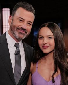 a man standing next to a woman in a purple dress and suit smiling at the camera