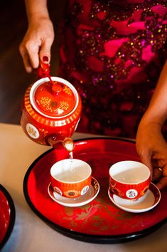 a woman pouring tea into two cups on top of a red saucer and plate