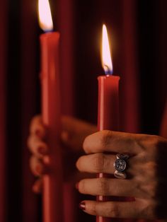 a woman's hand holding two candles in front of red curtains with one candle lit