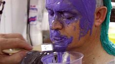 a man with purple paint on his face is painting another person's face in blue