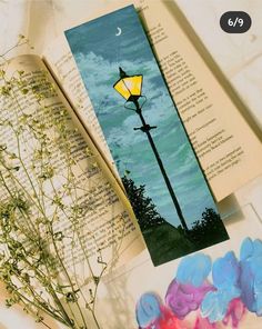 an open book with a lamp post and flowers on it next to some watercolor paints