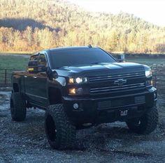 a large black truck parked on top of a gravel field next to a forest covered hillside