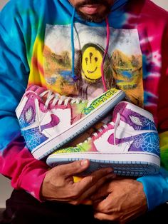 a man in a tie - dye hoodie holds up his sneakers with the image of a smiley face on them