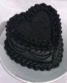 a black heart shaped cake with writing on the top and bottom is sitting on a white plate