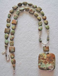 a long necklace with green and brown beads on white paper next to a metal hook