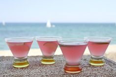 four glasses filled with pink liquid sitting on top of a table near the ocean and sailboats