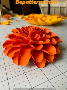 an orange paper flower sitting on top of a white tiled floor next to yellow flowers