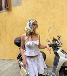 a woman in a white dress standing next to a scooter with a bandana on her head