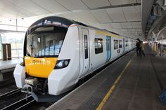 a yellow and white train pulling into a station with people standing on the platform next to it