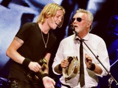 two men standing next to each other while holding guitars and singing into microphones on stage