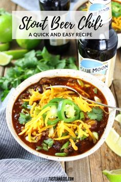 a white bowl filled with chili, cheese and green onions next to two bottles of beer