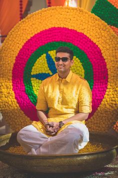 a man sitting on top of a wooden bowl filled with yellow and red flowers in front of a large circular object