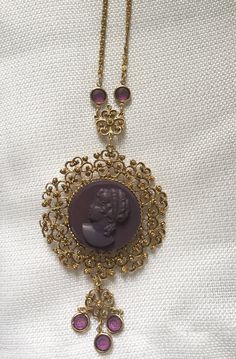 "Gorgeous 1950's statement watch pendant/necklace. Large gold plated medallion features a purple high relief carved profile cameo. Dangling down and attached to the chain are 7 round faux amethysts set in bezels. Lovely specialty chain has a large spring ring closure. The back has a Medana 17 Jewel mechanical wind watch, working well and winds easy. Necklace is 32\" long The pendant is 2 1/2\" in diameter My husband Alan & I had a brick & mortar store in Center City Philadelphia, on Jewe Brass Medallion Costume Jewelry, Vintage Costume Jewelry Medallion Necklace, Vintage Yellow Gold Cameo Necklace, Vintage Medallion Costume Jewelry Necklace, Vintage Yellow Gold Cabochon Necklace, Ornate Medallion Necklace With Cabochon, Purple Filigree Pendant Jewelry, Ornate Medallion Cabochon Necklace, Costume Jewelry Medallion With Vintage Charm