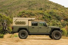 a green jeep parked on top of a dirt road next to a hill with trees