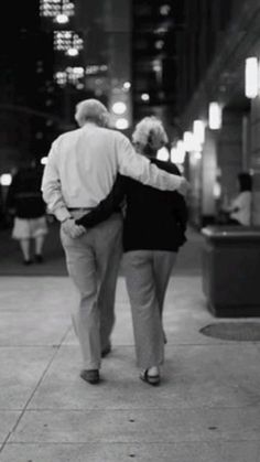 an older couple walking down the sidewalk in front of a building at night, back to back