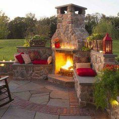 an outdoor fireplace is lit up in the evening with red pillows and candles on it