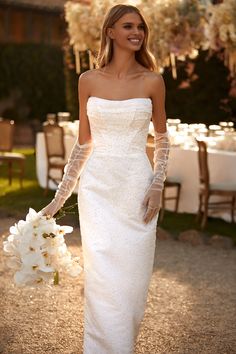 a woman in a white wedding dress holding a bouquet and smiling at the camera with her hands behind her back