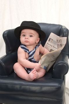 a baby sitting in a chair with a hat on and reading a newspaper while wearing a tie