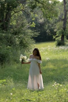 a woman in a white dress is standing in the grass and holding a bouquet of flowers