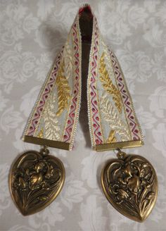 two gold heart shaped pendants are on a white tablecloth with floral designs and leaves