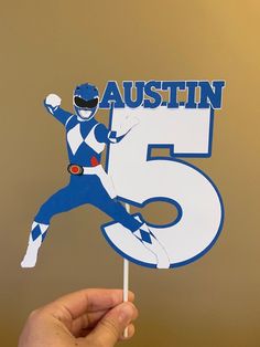 a hand holding up a blue and white cake topper with the number five on it