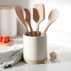 several wooden spoons in a white cup on a counter next to garlic and tomatoes