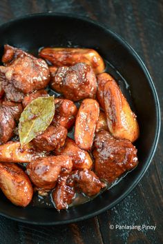 a black bowl filled with meat covered in sauce on top of a wooden table next to a green leaf