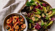 a bowl of shrimp next to a plate of salad on a white cloth with a fork