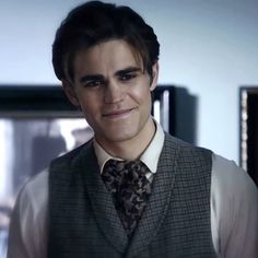 a young man wearing a vest and tie in a dark room with a window behind him