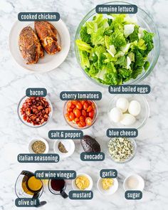 the ingredients for chicken salad laid out on a white marble counter top, including lettuce, eggs, bacon, cherry tomatoes, and dressing