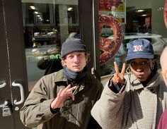 two people standing in front of a doughnut shop making the peace sign with their hands