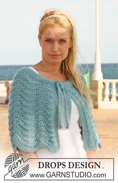 a woman standing on the beach wearing a blue knitted shawl and white top