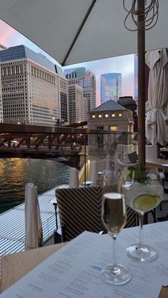 two glasses of wine sitting on top of a table next to a river with buildings in the background