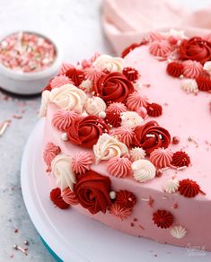 a heart shaped cake with pink frosting and red flowers on the top, surrounded by confetti sprinkles