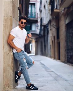 a man leaning up against a wall in an alleyway wearing sunglasses and ripped jeans